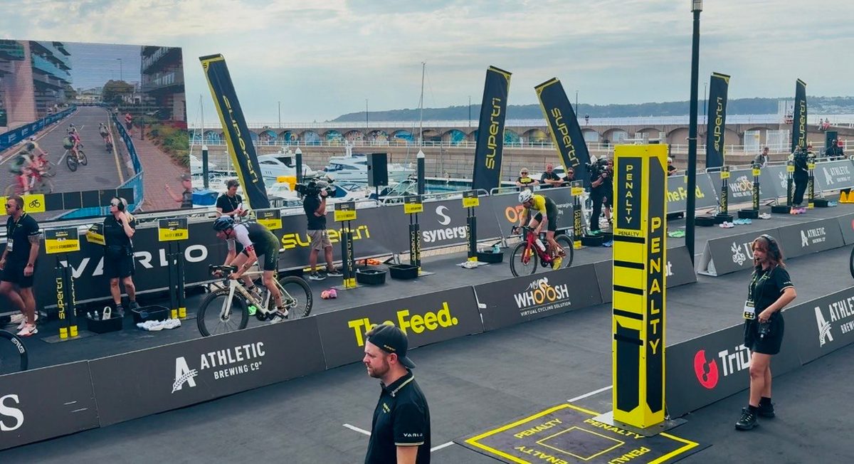 Riders leaving the transition area past a penalty box on the harbour at St Helier